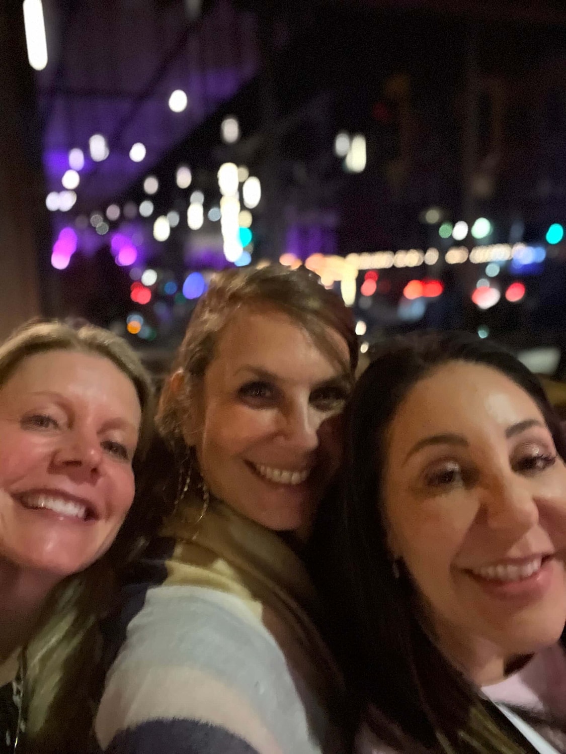 Three women smiling in urban night setting.