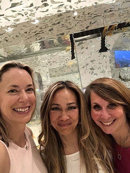 Three women smiling together in a reflective environment.